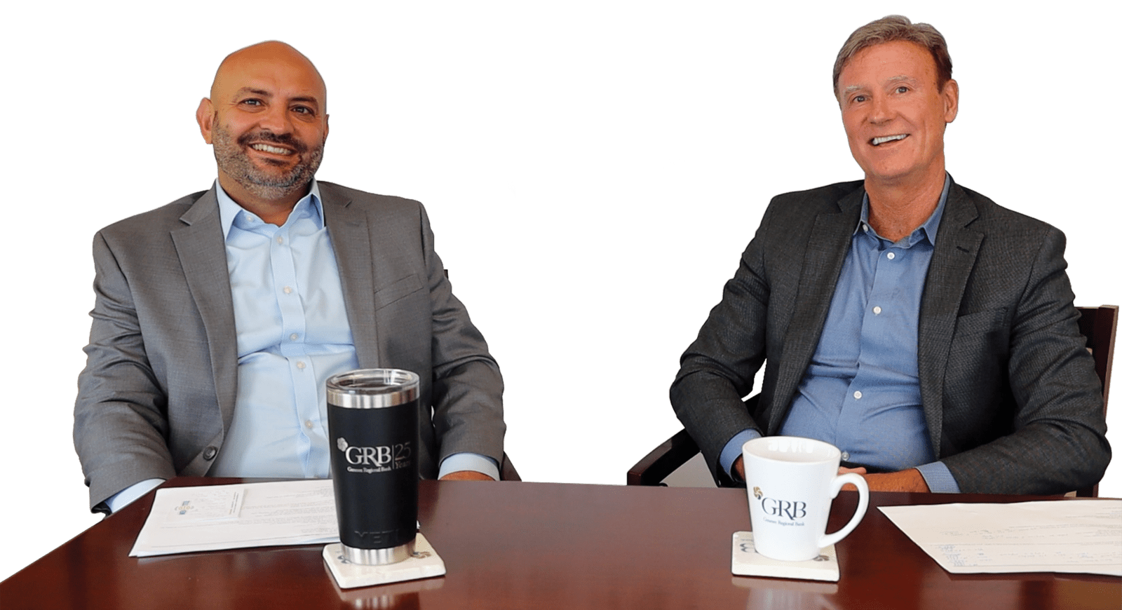 Joe Leone (left) and Mike Pulver (right) sit at a wooden table with GRB branded coffee mugs and papers in front of them.