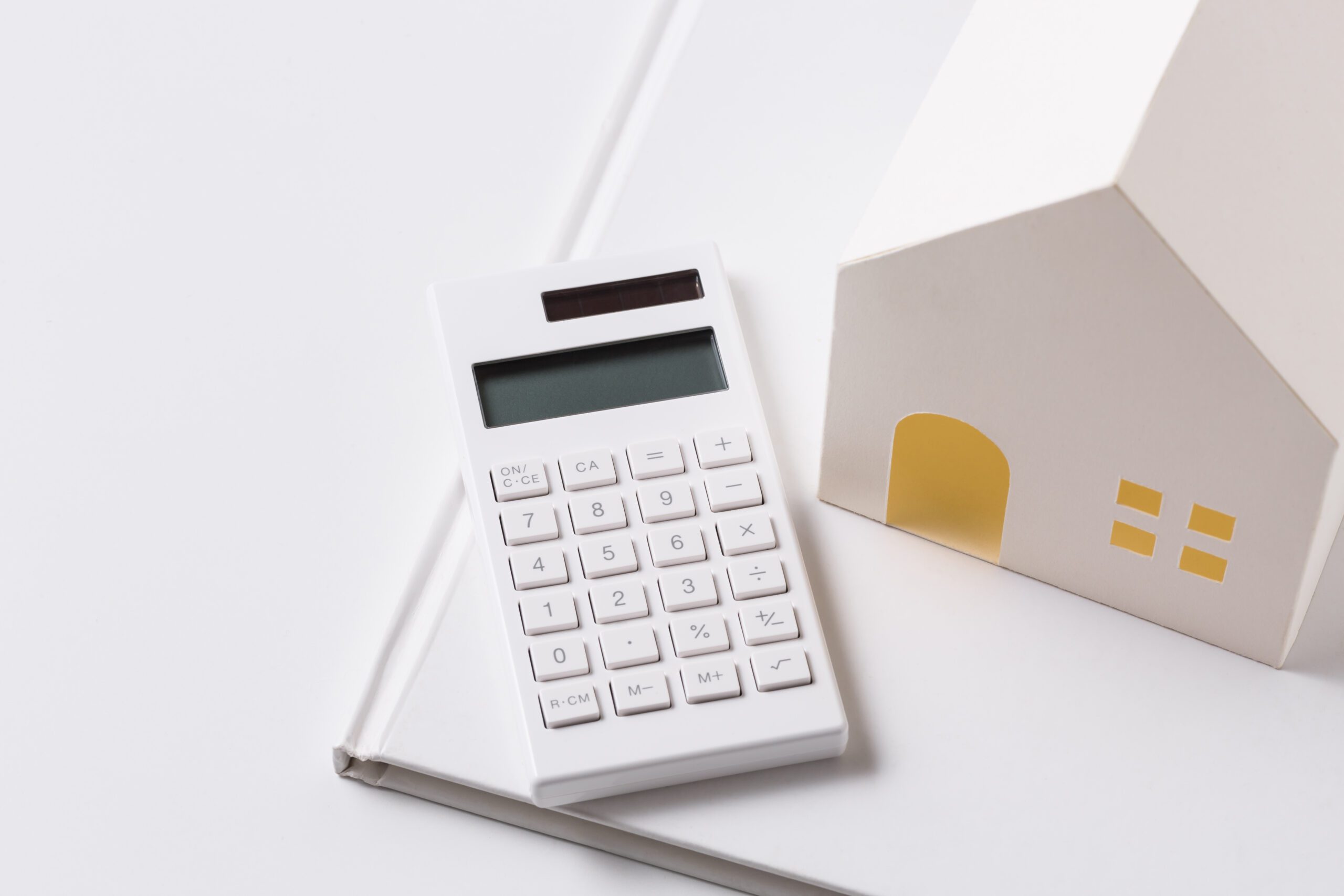 White 3D model of a house sitting on top of a white book on a white desk with a white calculator resting next to the model home.