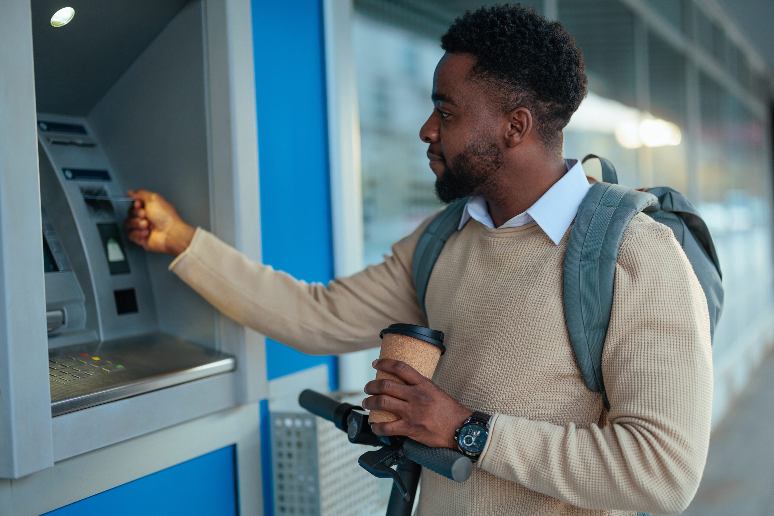 A focused, male adult is seen inserting a debit card into an ATM machine while holding a coffee cup and wearing a backpack.