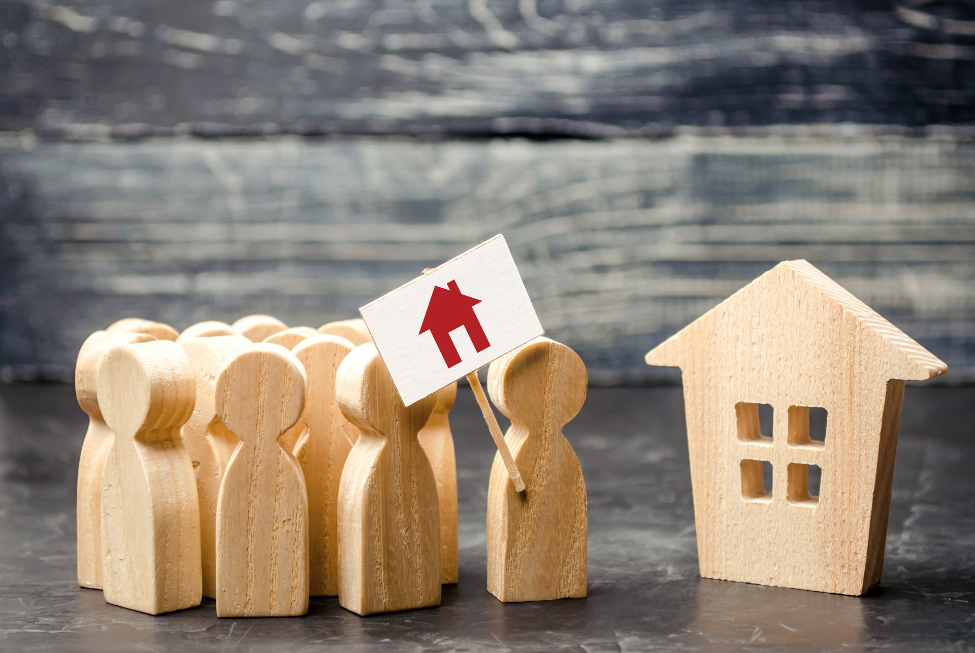 A large group of wooden cutout figures line up to see one house. Represents shrinking housing inventory.