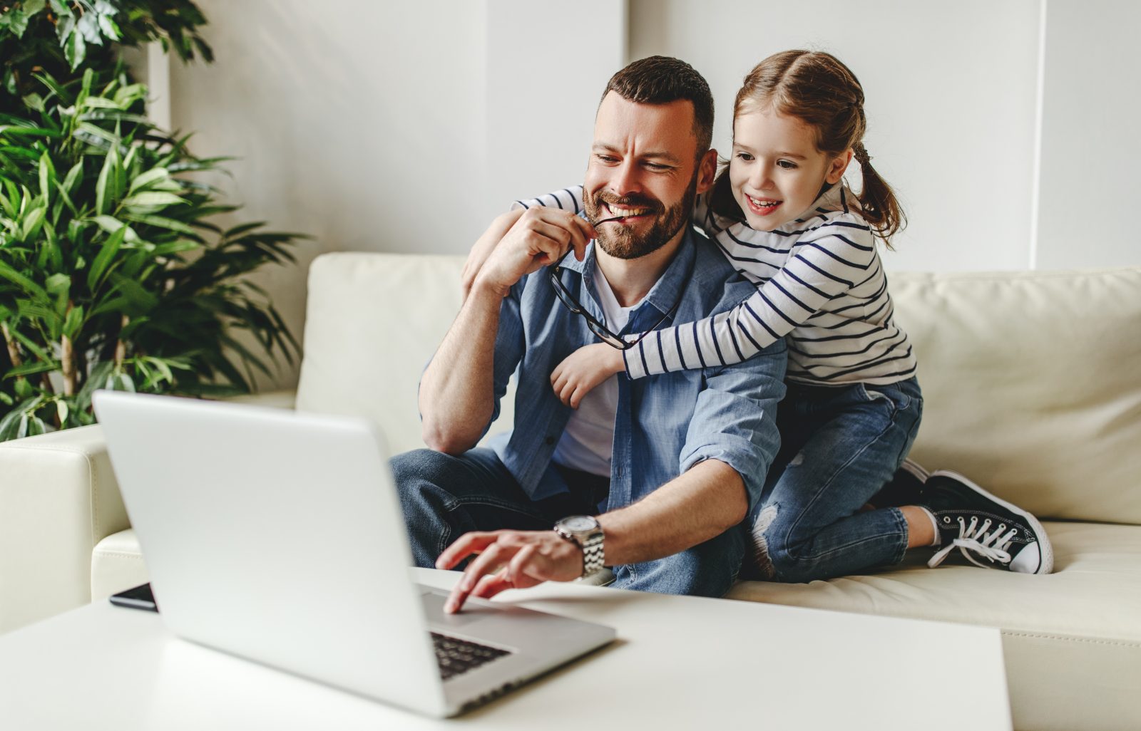 Man working on his laptop on a coffee table while his daughter sits next to him on the couch and hugs him.